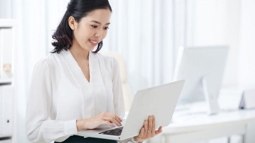 Standing woman in white blouse types on white laptop in an office of white furniture in Singapore