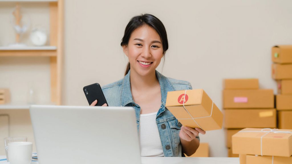 Young female small business owner smiling holding a package and phone behind laptop in Singapore