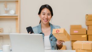 Young female small business owner holding a package and phone behind laptop in Singapore