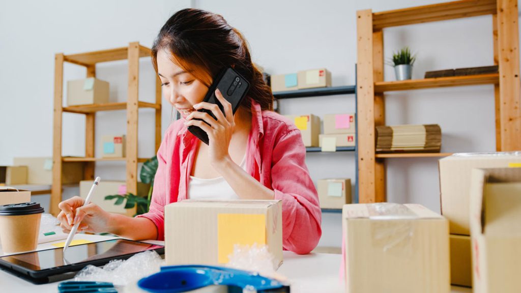 Young female start-up business owner in Singapore talking on the phone while using a stylus tablet with packages on the table