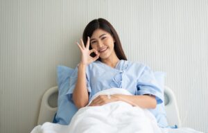 Asian woman in hospital bed flashing an Ok sign after securing a quick loan for medical purposes.